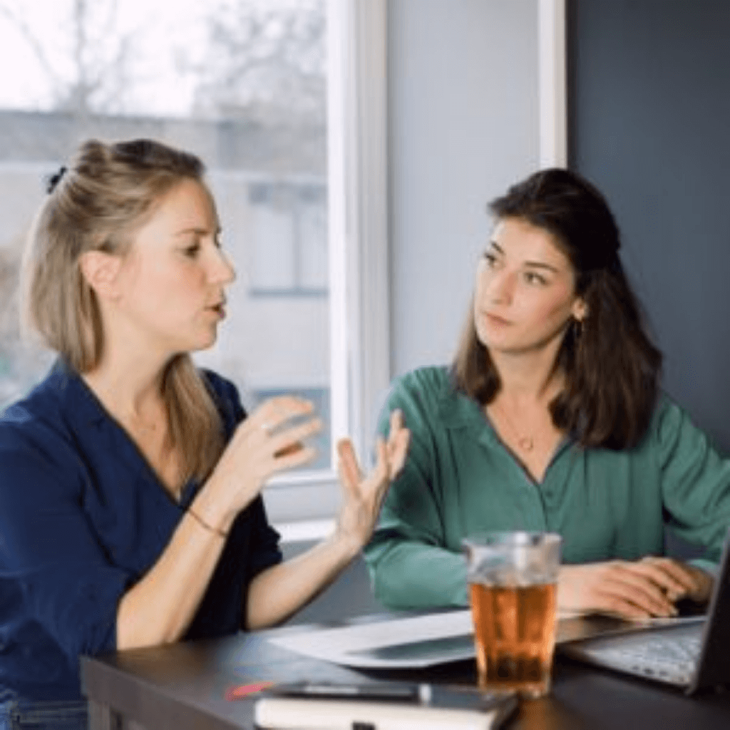 A woman sitting at a table with a cup of tea is explaining something to another woman next to her, who is looking at her. The woman who is explaining is clearly using her hands to emphasize her explanation.