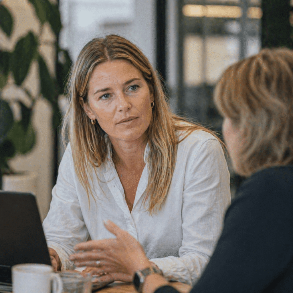 2 women who have a conversation in a business setting. The women in the middle of the picture is listening to the other women on the right side of the picture. They have a cup of coffee and a glass of water in front of them.
