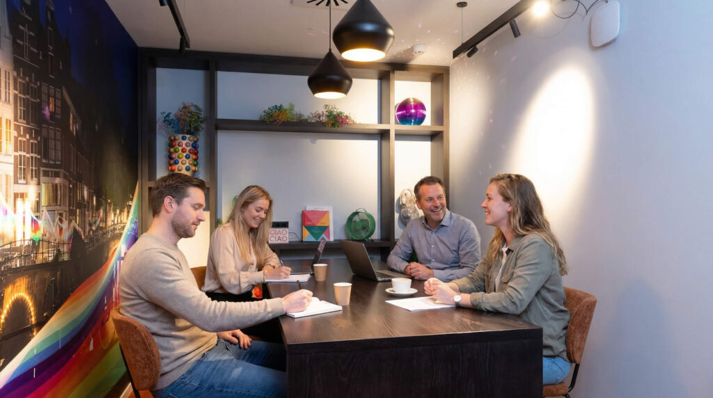 4 people are sitting in a meeting room making notes on paper and on their laptops. Theey are sitting on brown chairs at a table. The meeting room has a very happy atmosphere with a lot of different colours.